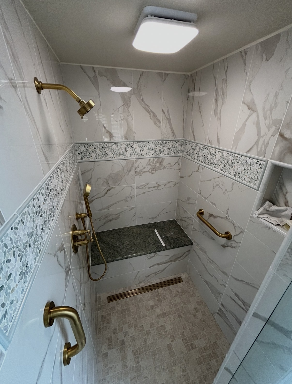 Completed marble walk-in shower with brass fixtures, mosaic tile band, and bench seat